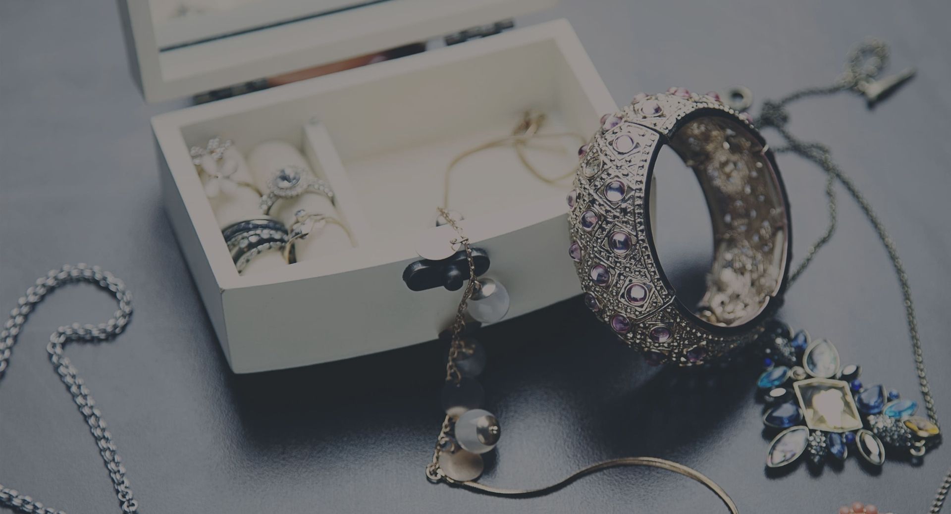 Offene Schmuckschatulle mit Ringen, Armreif und Ketten auf dunkler Oberfläche.